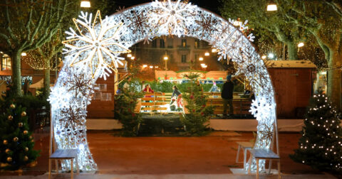 Arche lumineuse en premier plan indiquant l'entrée du Village de Noël à Villefranche. En fond, des personnes s'amusent sur la patinoire.